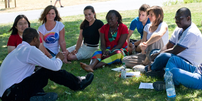 Eine Gruppe Studierender hört auf einer Wiese sitzend einem Dozenten zu