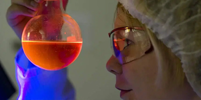 Frau mit Schutzbrille blickt in Kolben mit leuchtender Flüssigkeit